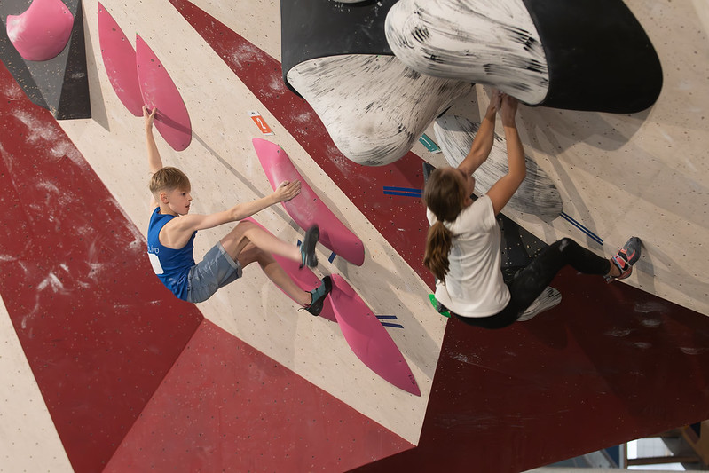 links jongen boulderend in blauw hempje aan roze grepen, rechts meisje boulderend in witte t-shirt aan marmere grepen